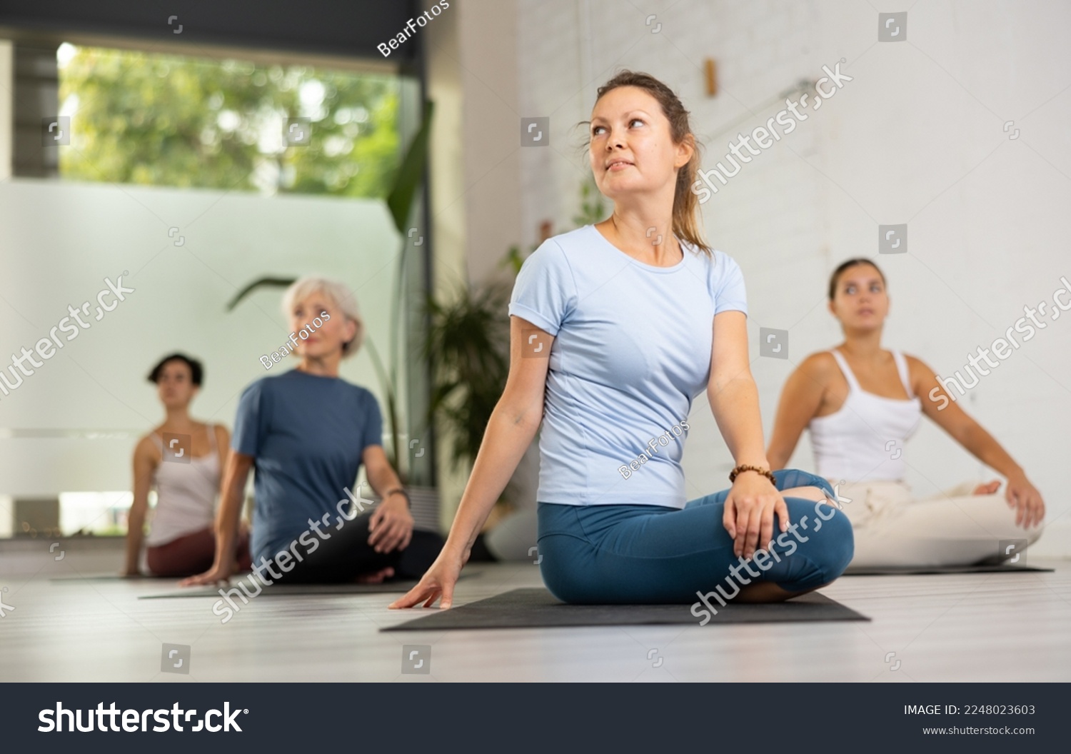 Relaxed middle-aged female performing Ardha Matsyendrasana position during group yoga training in fitness club #2248023603
