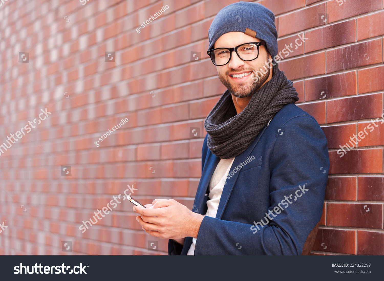 Typing text message. Side view of handsome young man in smart casual wear holding mobile phone while leaning at the brick wall #224822299
