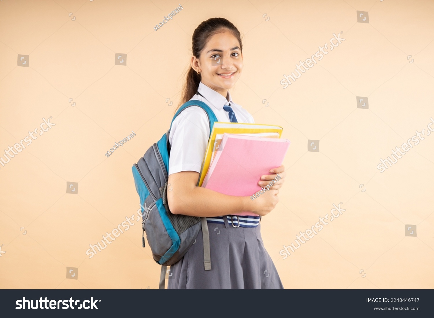 Happy Indian student modern schoolgirl wearing school uniform holding books and bag standing isolated over beige background, Studio shot, Education concept. #2248446747