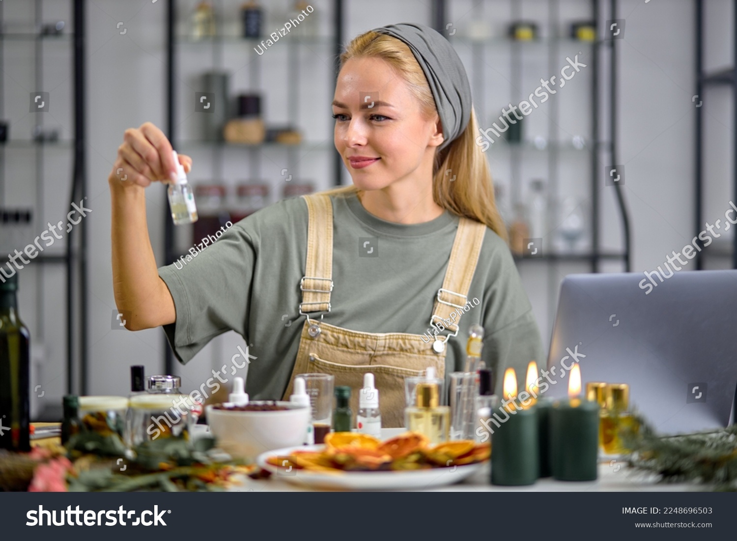 Woman going to mix ingredients to make perfume with many bottles of essential oil. sit behind table using laptop, indoors in cozy room alone. blonde female perfumer creates fragrance #2248696503