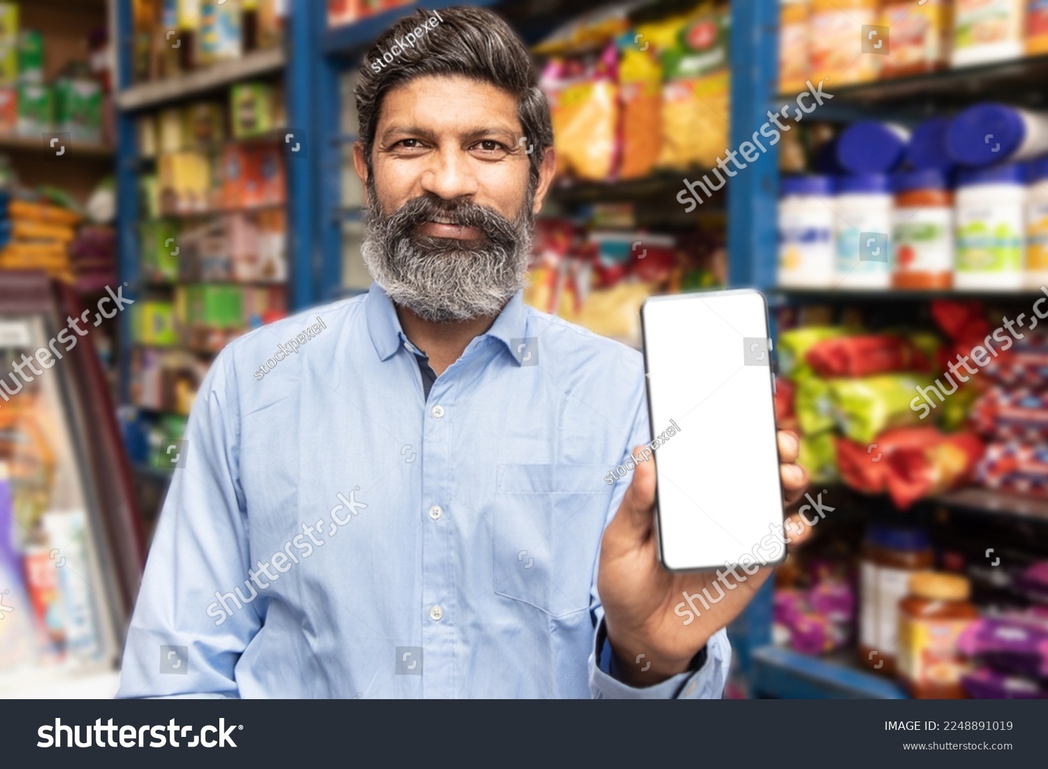 Portrait of happy mature beard Indian man showing smart phone with blank display screen to put advertisement at grocery shop or supermarket, Closeup. #2248891019