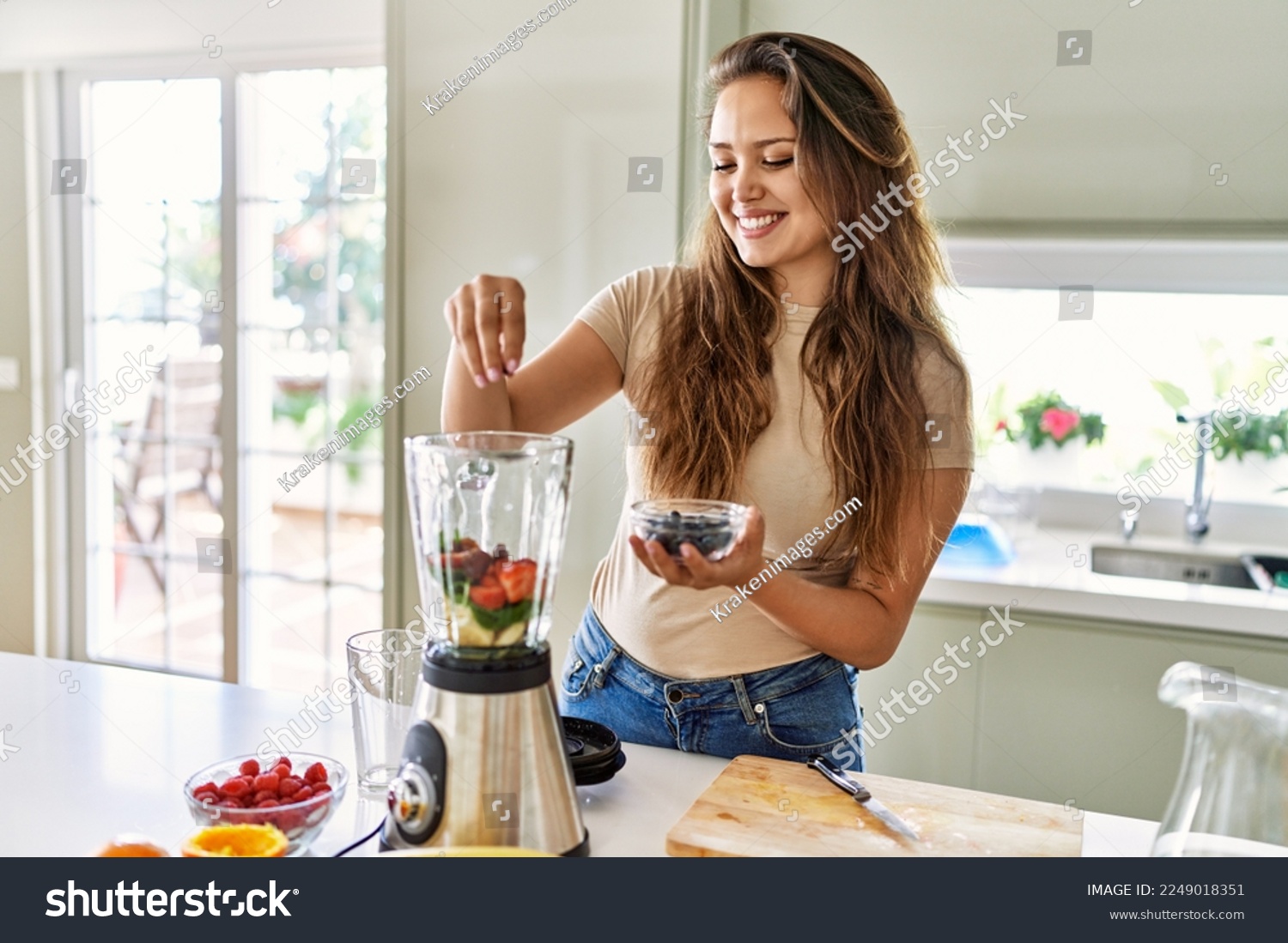 Young beautiful hispanic woman preparing vegetable smoothie with blender at the kitchen #2249018351