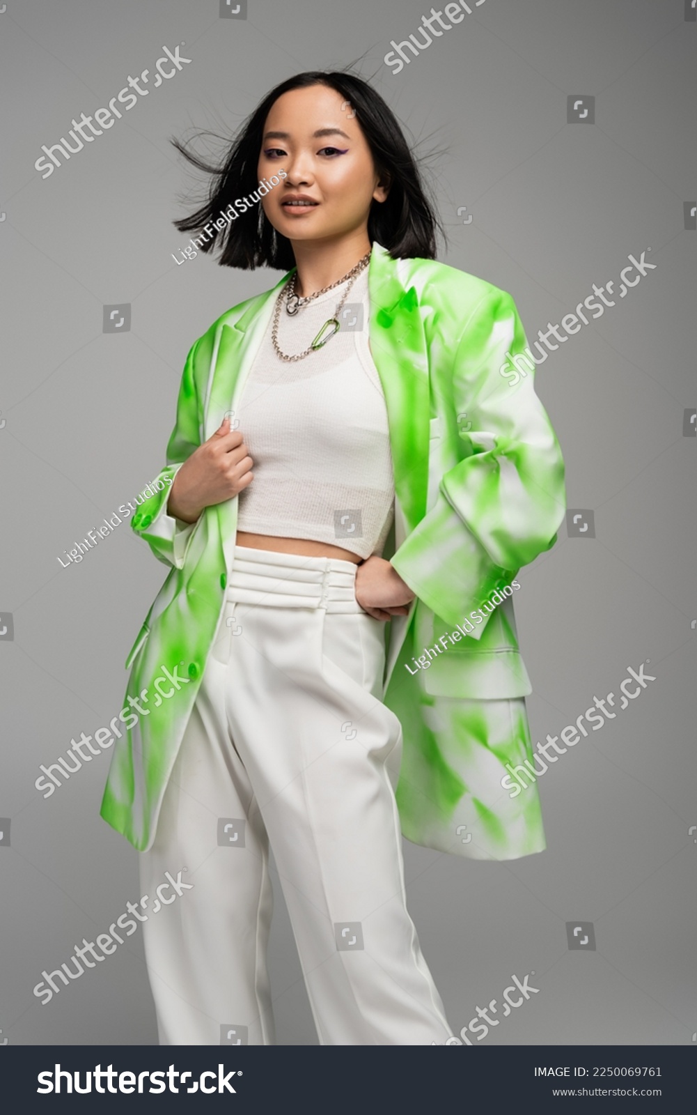 trendy asian woman in green and white blazer standing with hand on hip isolated on grey #2250069761