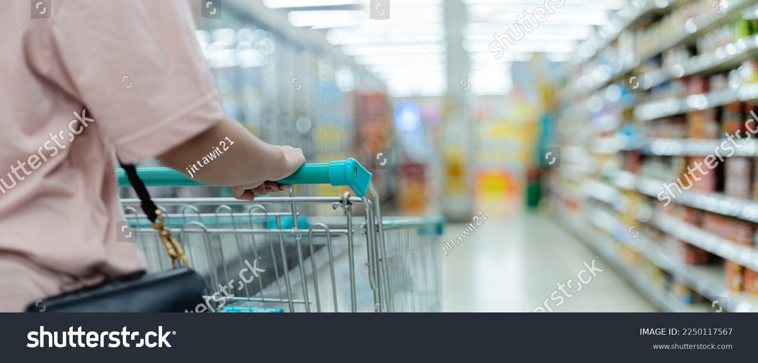 Woman doing grocery shopping at the supermarket, pushing a shopping cart. Shopping cart in supermarket aisle, copy space. Closeup of a woman shopper with a modern shopping cart at supermarket #2250117567