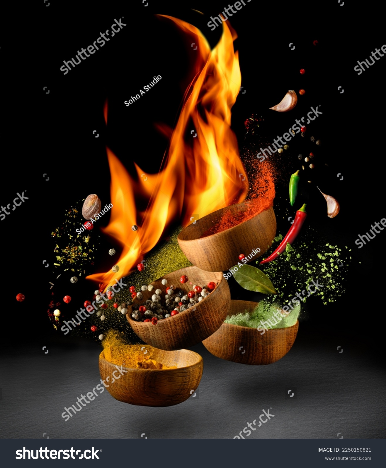 Hot spices and chili peppers in wooden bowls flying over black background and fire. Spices and seasonings powder with flame splash. Freeze motion photo. #2250150821