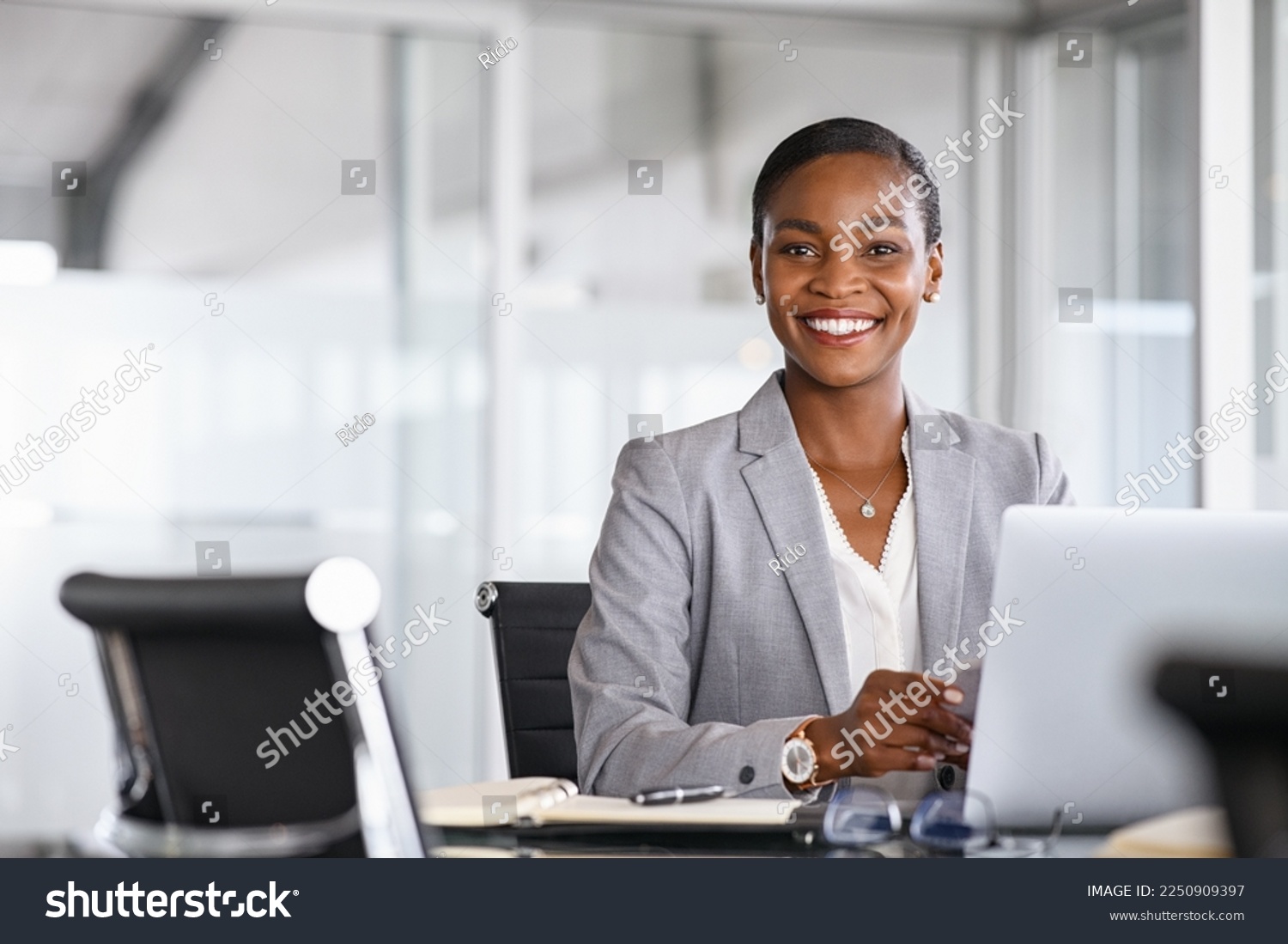 Portrait of a cheerful businesswoman sitting at desk in modern office and looking at camera. Smiling african american executive using laptop while working from office with copy space. #2250909397