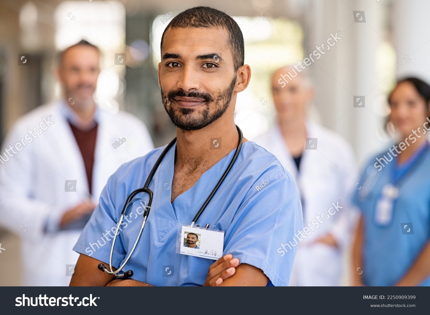 Portrait of smiling middle eastern man nurse with stethoscope looking at camera. Young doctor smiling while standing in hospital corridor with health care team in background. Successful indian surgeon #2250909399