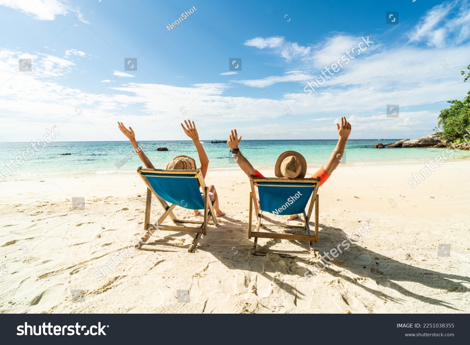 Two happy people having fun on the beach, sitting on blue sunbed with hands raised up, spending leisure time together. Summer holidays concept. Tourism. Travelers.
 #2251038355