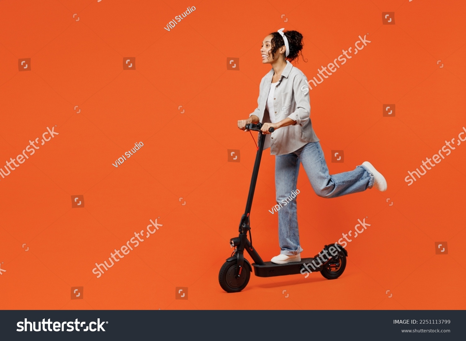 Full body young happy fun woman of African American ethnicity she wears grey shirt headband riding e-scooter look aside isolated on plain orange background studio portrait. People lifestyle concept #2251113799