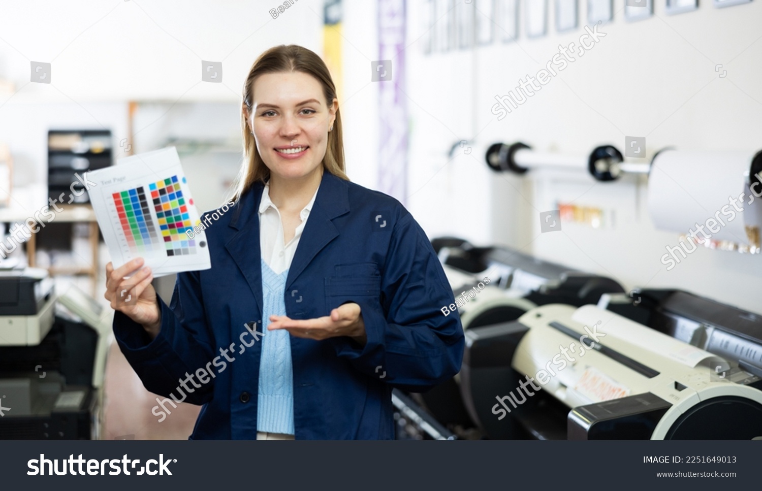 Portrait of positive woman printing office worker holding colour test page for printer and making presenting gesture. #2251649013