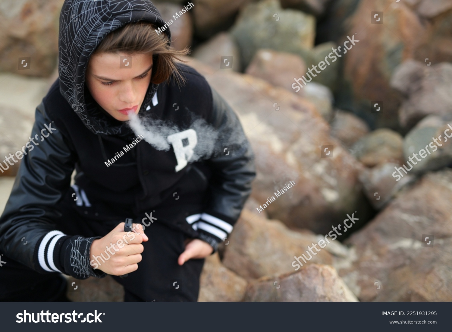 a guy of thirteen years old, holds a vape in his hands. A teenager smokes a disposable electronic cigarette while sitting by the sea #2251931295