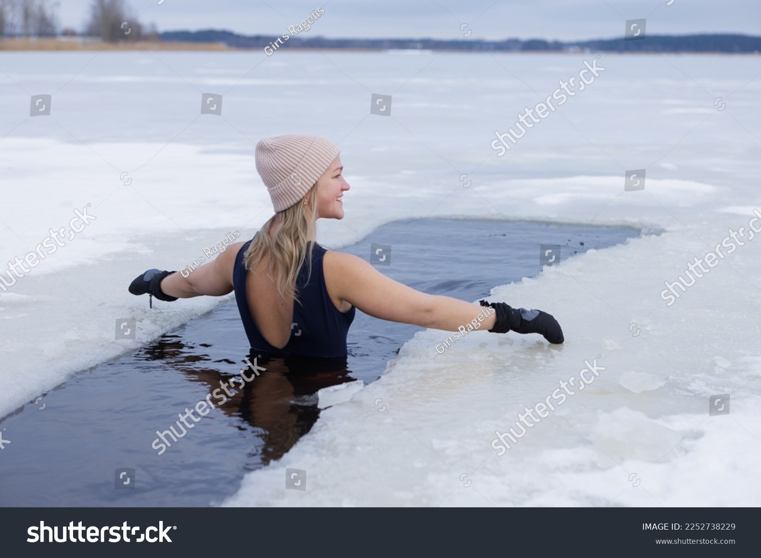 Winter swimming. Woman in frozen lake ice hole. Swimmers wellness in icy water. How to swim in cold water. Beautiful young female in zen meditation. Gray hat and gloves swimming clothes. Nature lake #2252738229