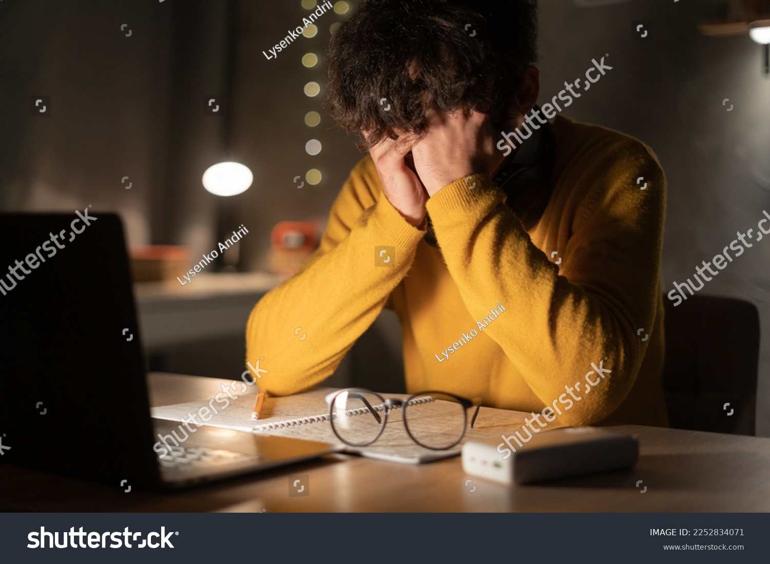 Tired and exhausted young man hides eyes with hands at laptop pc late in the evening. Depressed male student sitting in front of computer screen at night. Concept of anxiety. Copy space #2252834071