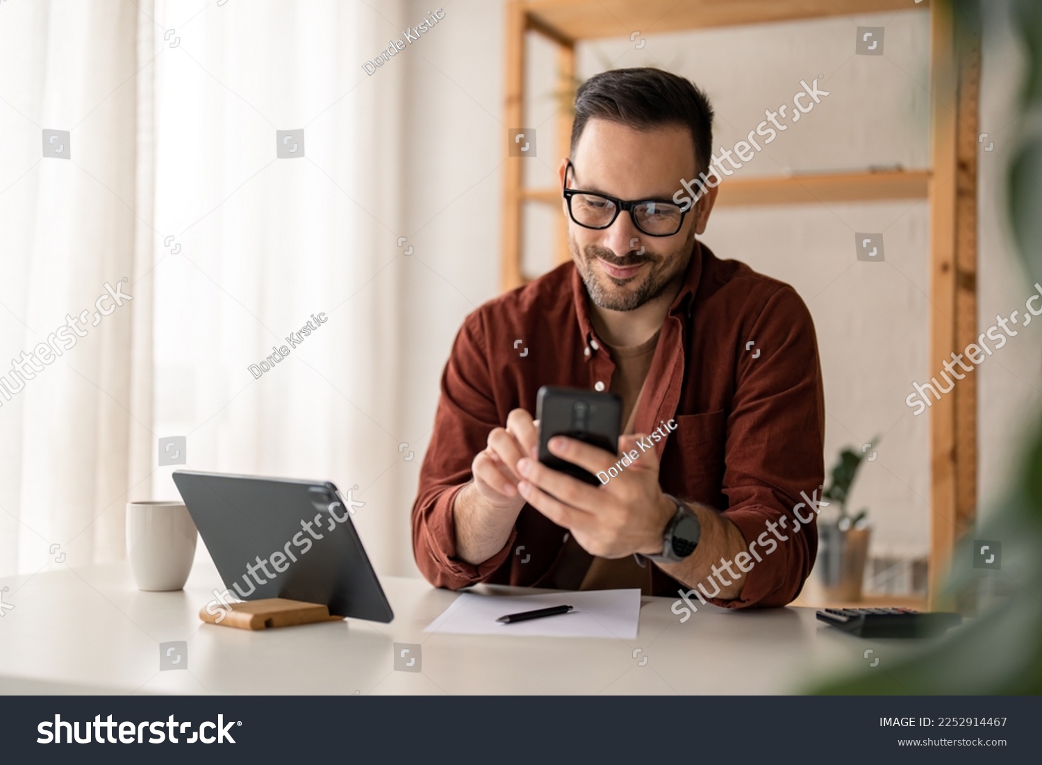 Happy smiling businessman wearing casual clothes and using modern smartphone in his home office during the day, typing, touching the screen, browsing the internet or writing text messages. #2252914467