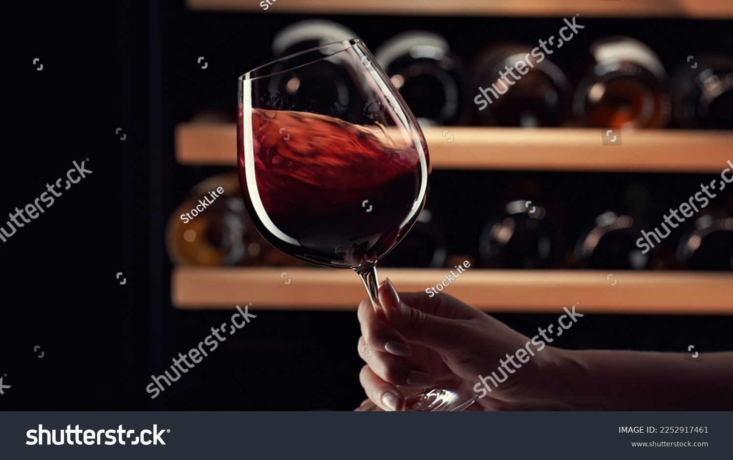 Close up female hand swirling red wine in wine glass. Wine expert tasting, rating and drinking wine, bottles in background. #2252917461