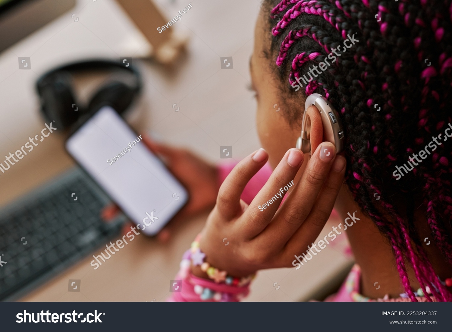 Close up of black teenage girl wearing hearing aid on ear and pressing button while using smartphone at home, copy space #2253204337