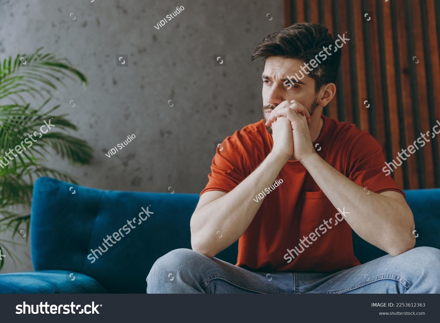Young minded sad man wear red t-shirt prop up chin look aside sit on blue sofa couch stay at home hotel flat rest relax spend free spare time in living room indoors grey wall. People lounge concept #2253612363