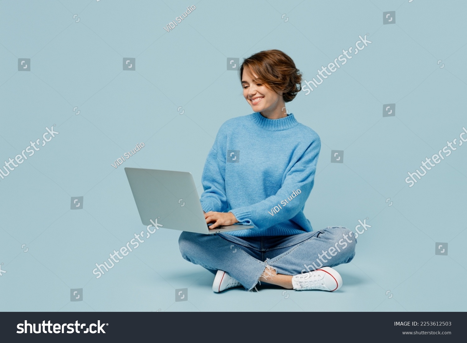 Full body young smiling happy fun cool IT woman wear knitted sweater hold use work on laptop pc computer isolated on plain pastel light blue cyan background studio portrait. People lifestyle concept #2253612503