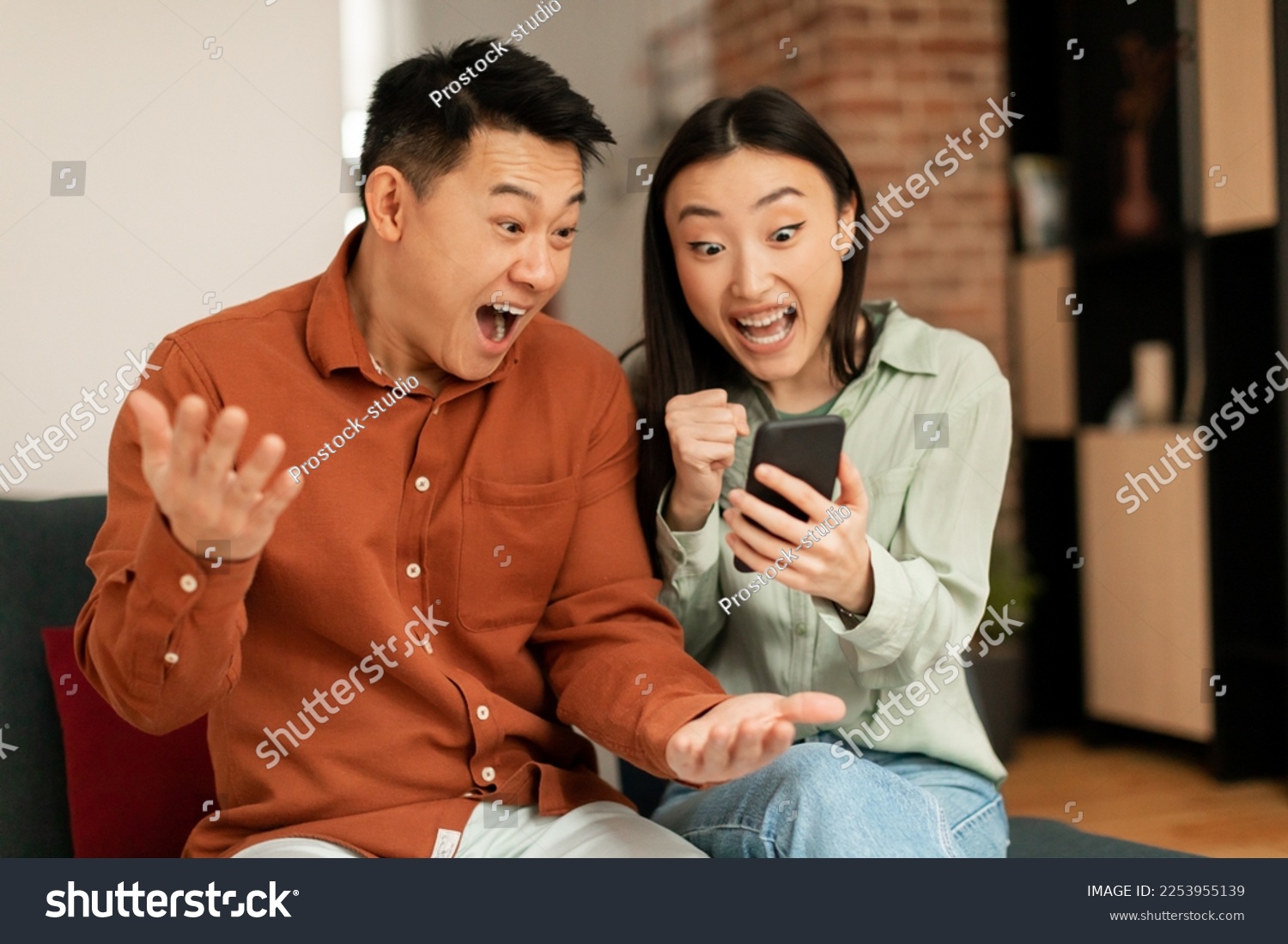 Portrait of overjoyed asian couple using smartphone and making winner sign gesture, shaking fists, sitting on couch at home, reading great news #2253955139