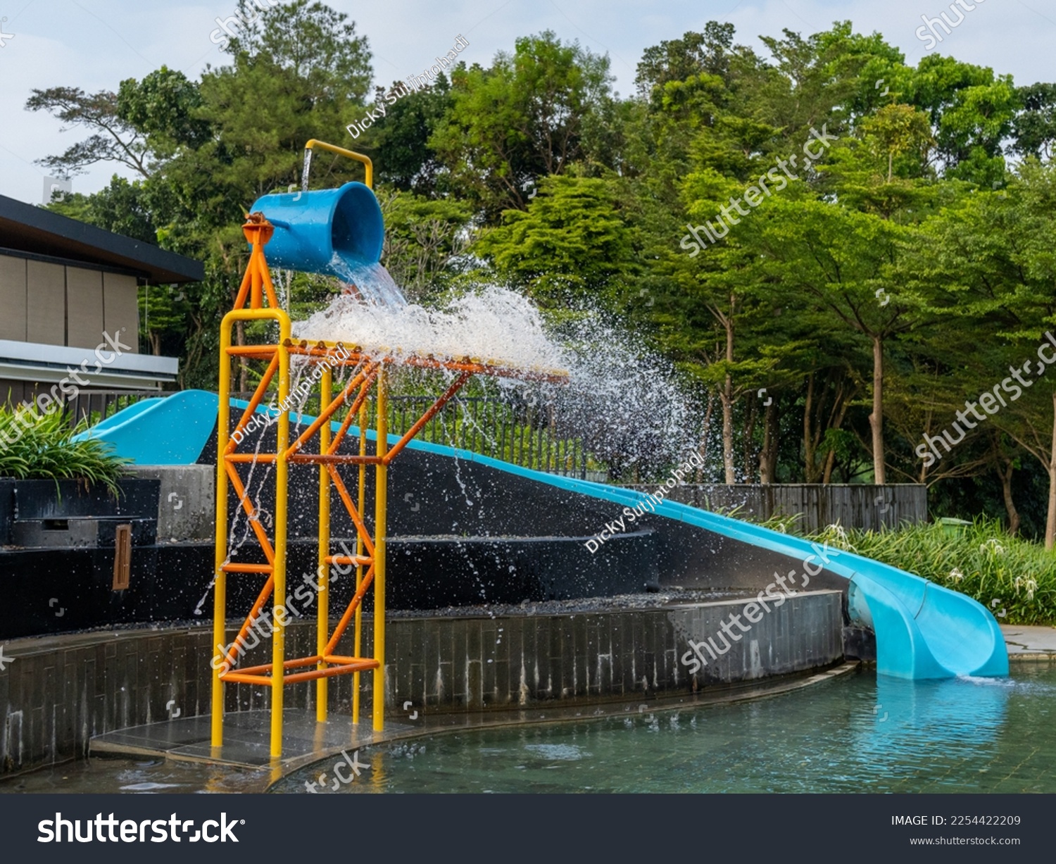 A blue water slide next to a water pouring large blue bucket on top of a colorful metal scaffolding at the side of a children pool. #2254422209