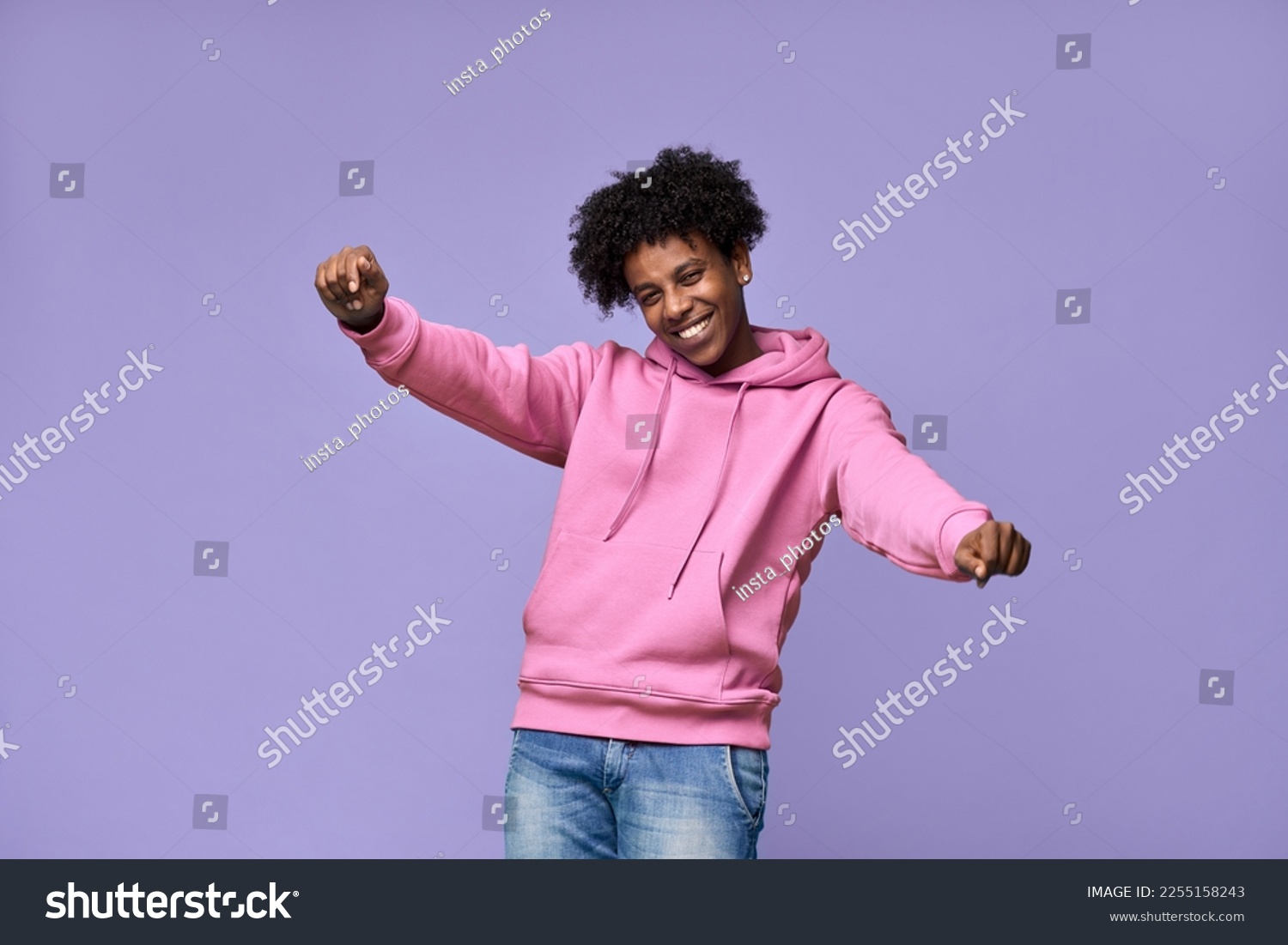 Young happy funky African American teen guy wearing pink hoodie having fun isolated on light purple background. Smiling cool ethnic generation z teenager student model dancing and moving. #2255158243