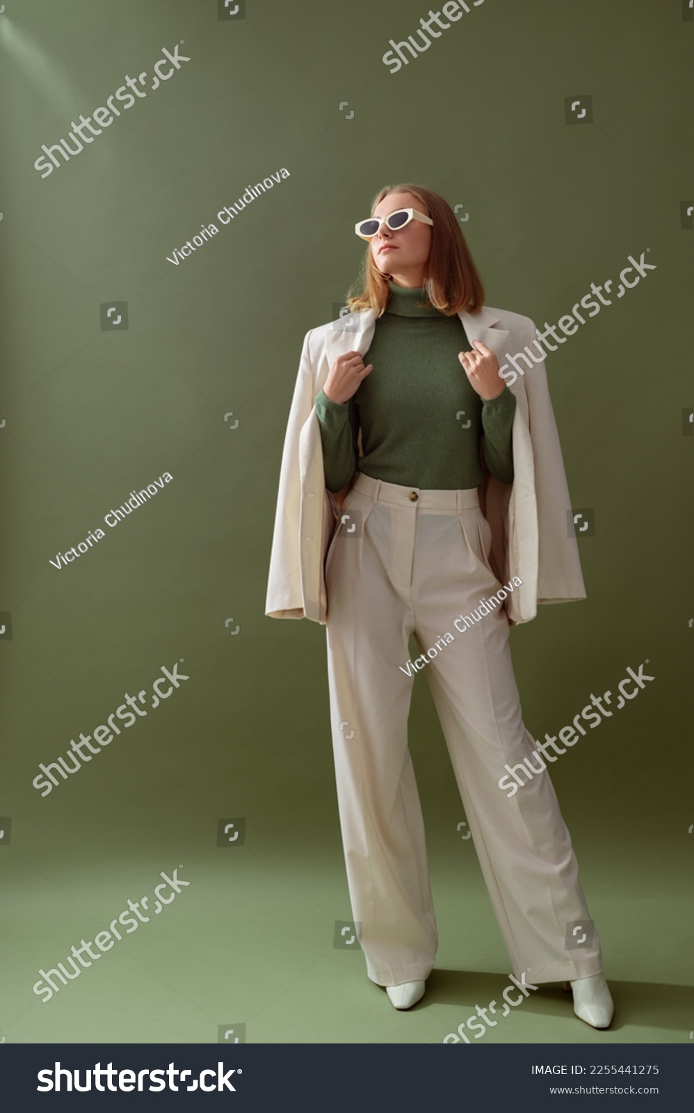 Fashionable confident woman wearing elegant white suit with blazer, wide leg trousers, cashmere turtleneck sweater, trendy sunglasses, posing on green background. Full-length studio fashion portrait #2255441275
