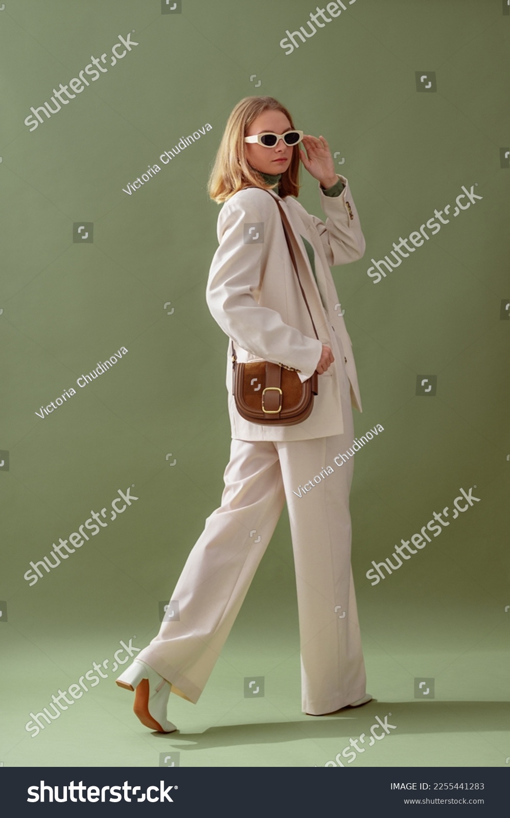 Fashionable confident woman wearing elegant white suit with blazer, wide leg trousers, trendy sunglasses, brown suede shoulder bag, posing on green background. Full-length studio fashion portrait #2255441283