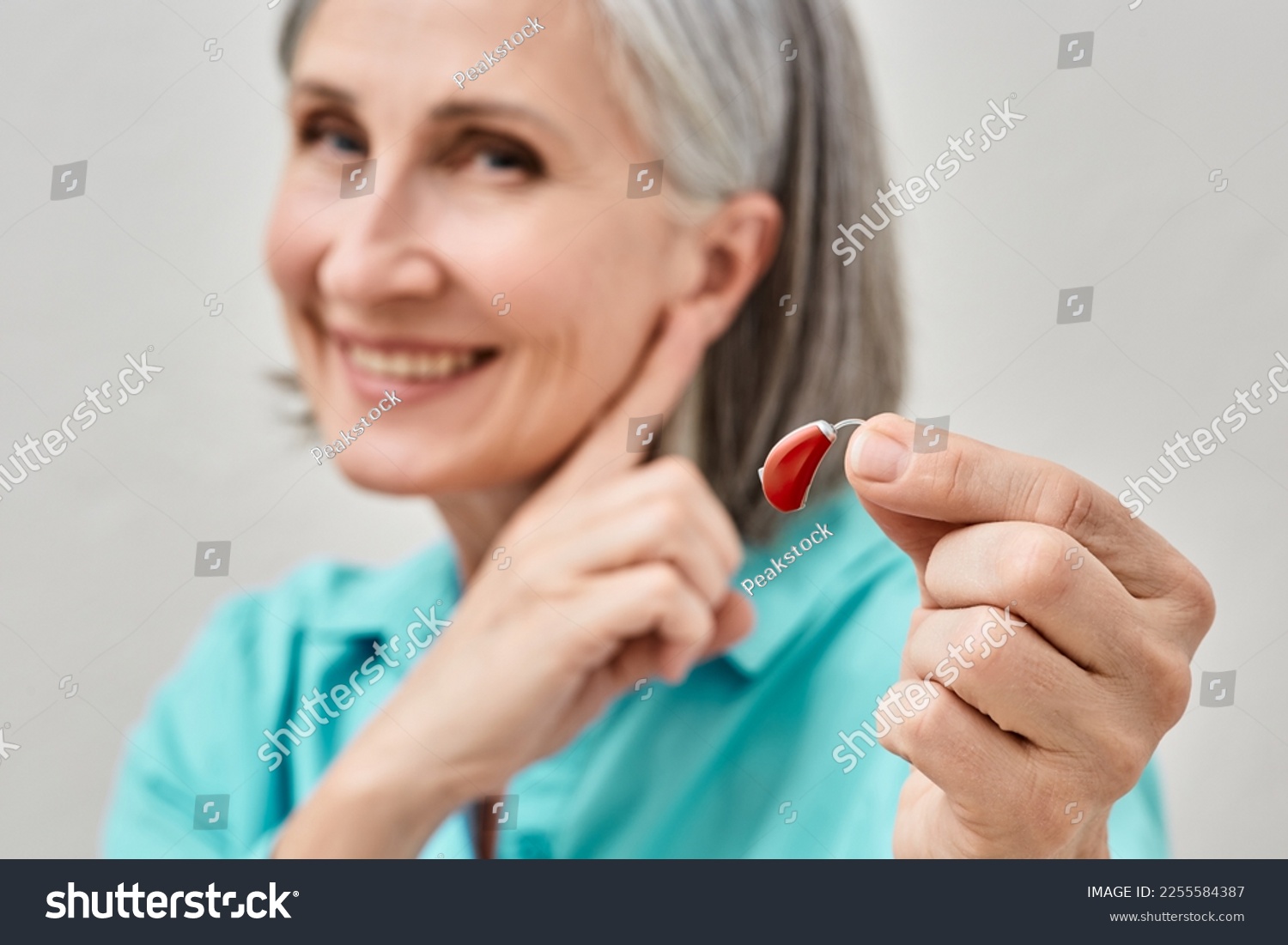 Senior woman holding hearing aid in hand on foreground and pointing finger at her ear, soft focus. Treatment of deafness in people with hearing aids #2255584387