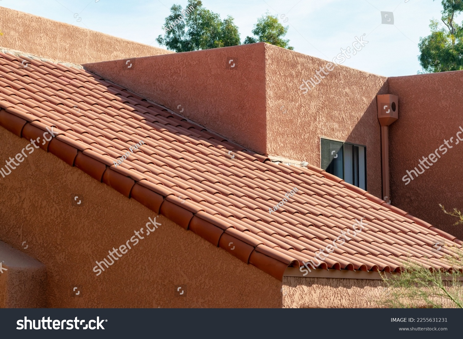 Long slanted adobe style roof with red tiles and orange stucco exterior with back and front yard trees in a neighborhood. In suburban area in late afternoon sun with desert design and facade. #2255631231