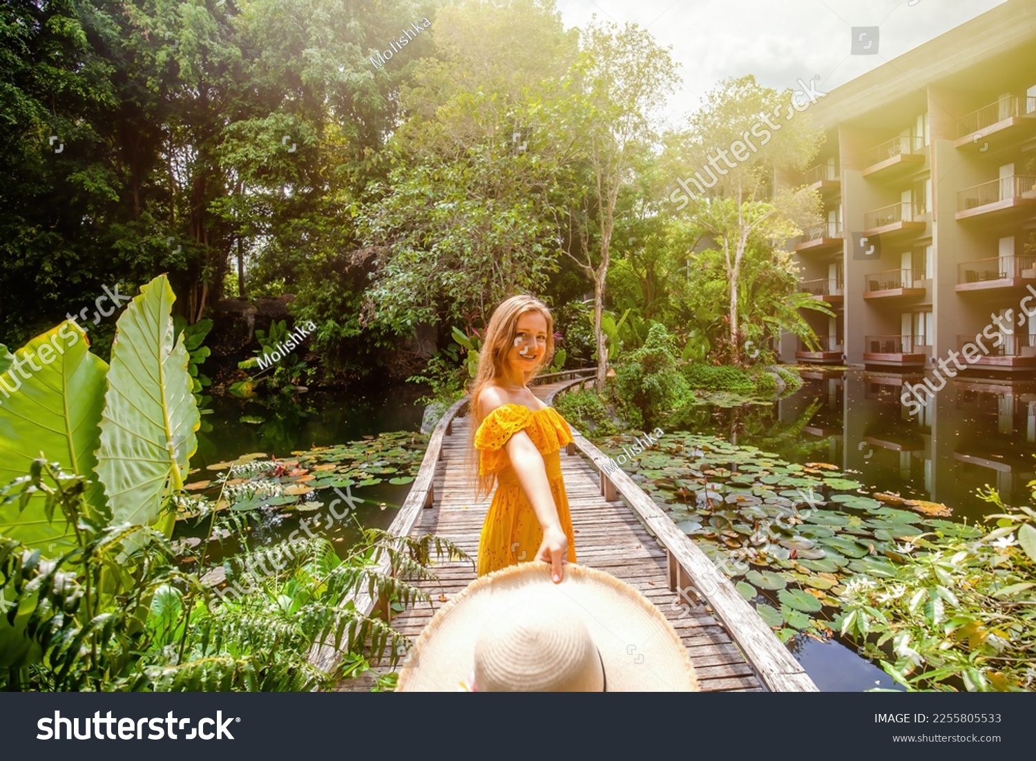 Happy woman in yellow dress dancing with straw hat in her hand, looking at camera. Summer vacation in resort. Female traveler on wooden bridge among tropical greenery and green pond with water lilies #2255805533
