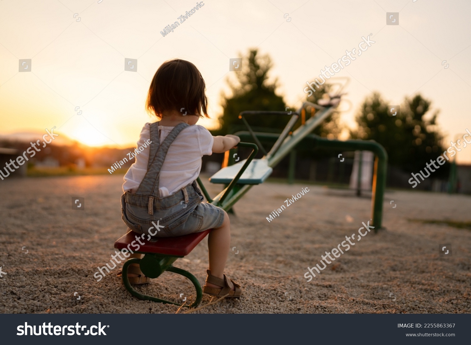 back view of one small caucasian toddler child sitting alone on the seesaw in park in sunset lonely with no friends copy space childhood growing up concept social issues rejected #2255863367