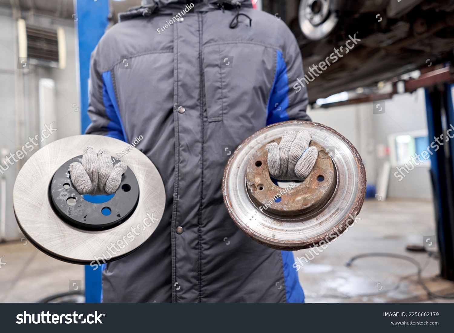 The mechanic holds old rusty brake disc and new disc. Change the old to new brake disc on car in a garage. Auto repair concept. #2256662179