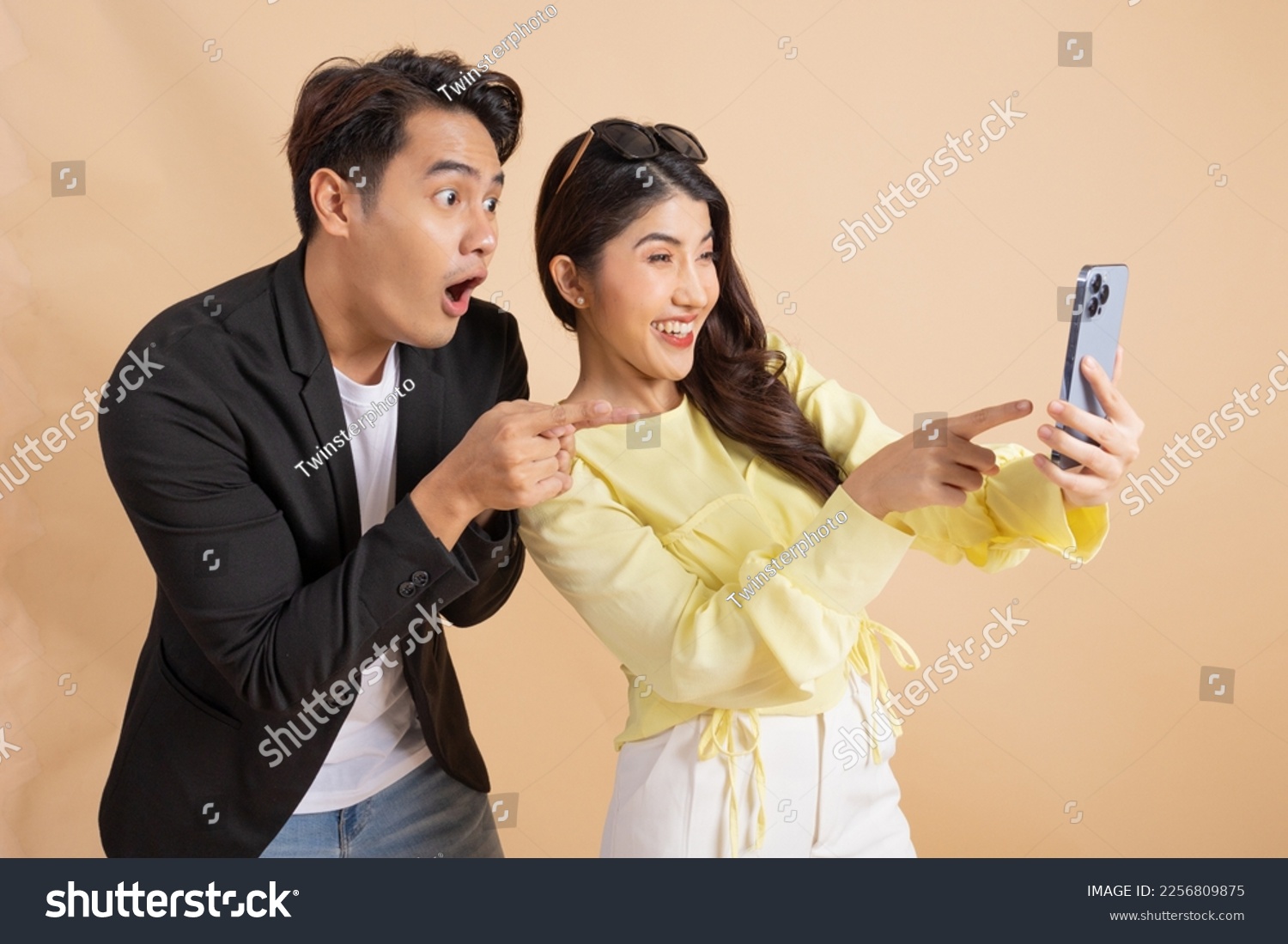 Shocked excited Asian businessman along with happy smiling woman pointing finger at smart phone over beige studio background. Man in white shirt and suit and woman in yellow top and white trousers. #2256809875