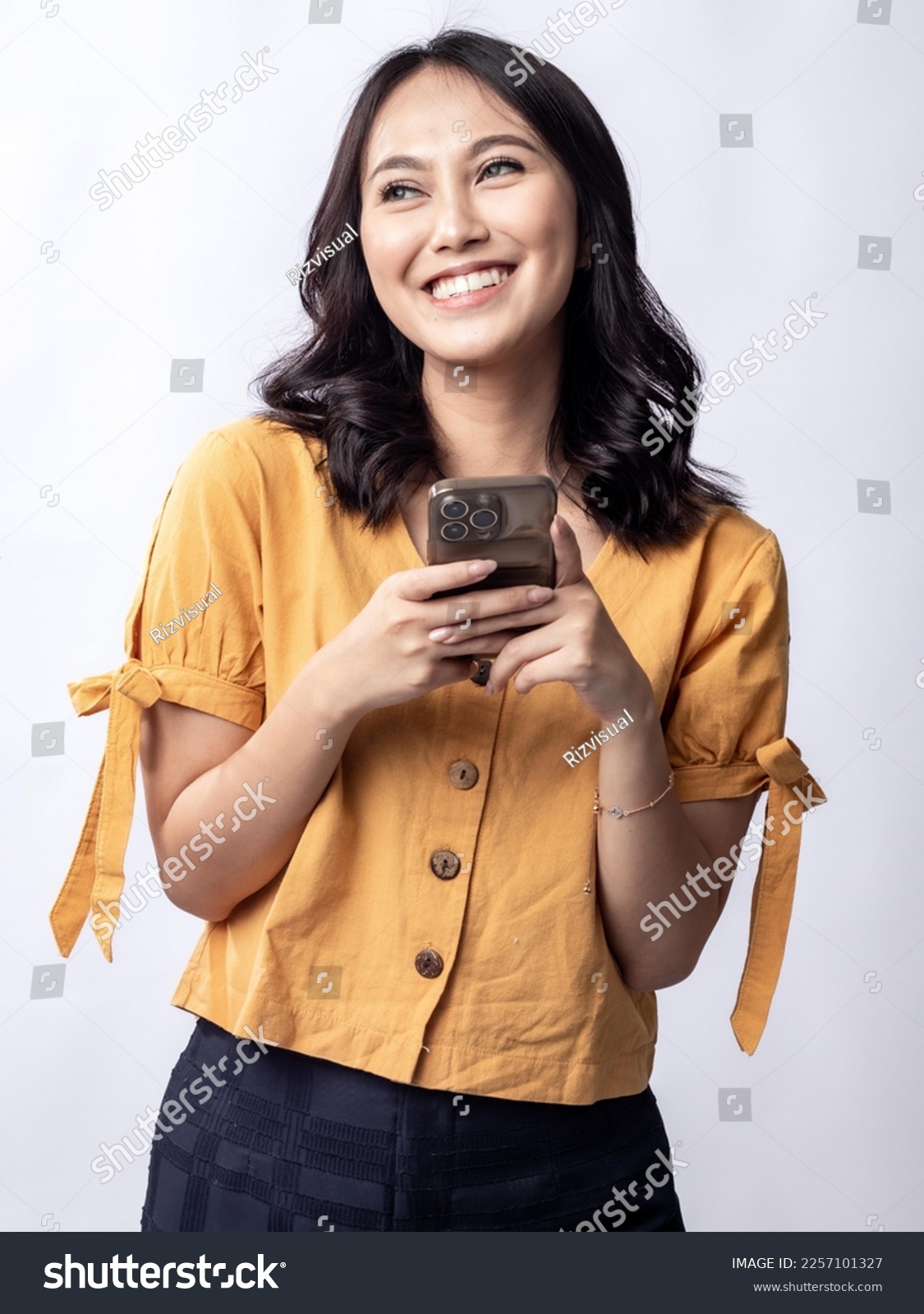 A studio portrait of a young beautiful Indonesian Asian woman in a casual yellow shirt looks happy while typing on her phone.  isolated on white background #2257101327