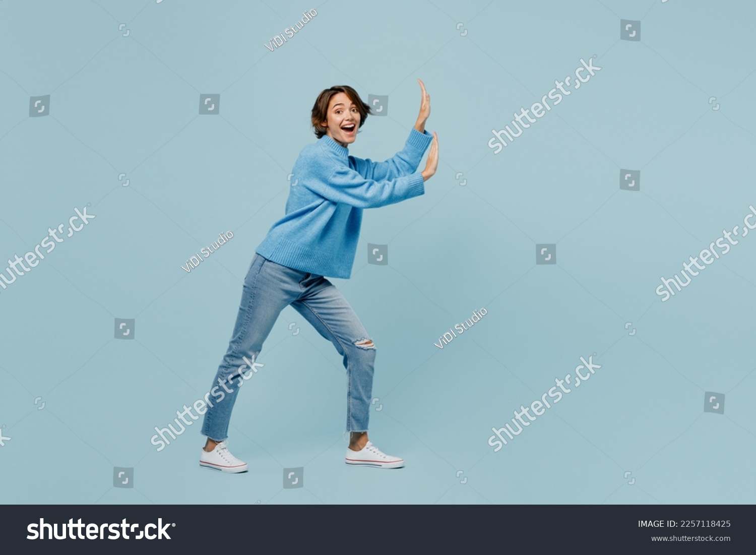 Full body young woman wear knitted sweater look camera push invisible object with copy space area mock up isolated on plain pastel light blue cyan background studio portrait. People lifestyle concept #2257118425