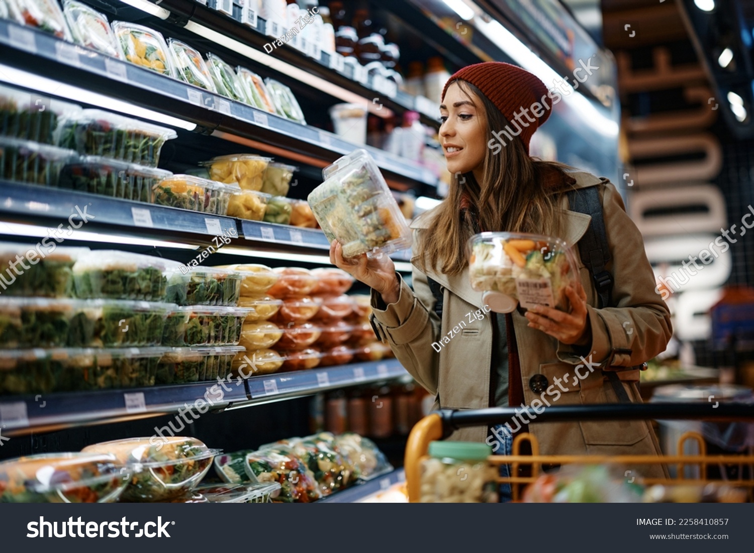 Smiling woman reading label on food package while buying groceries from refrigerated section in supermarket. #2258410857