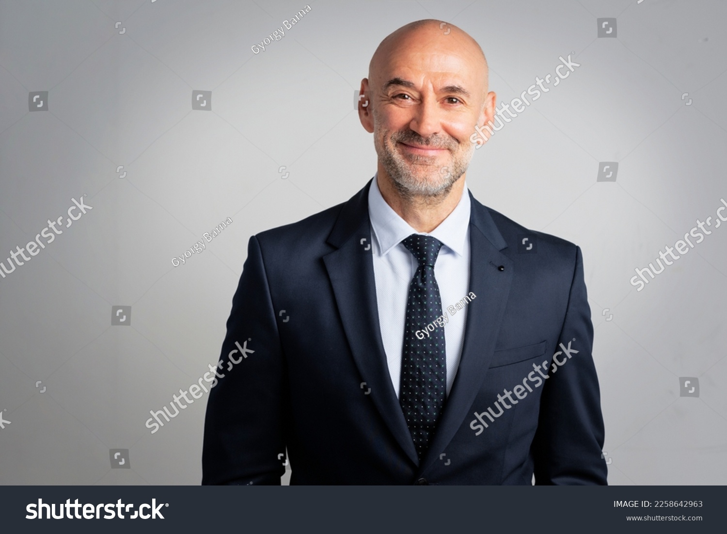 Portrait of caucasian business man looking at camera and smiling. Confident mature male professional is in suit. He is against gray background. #2258642963