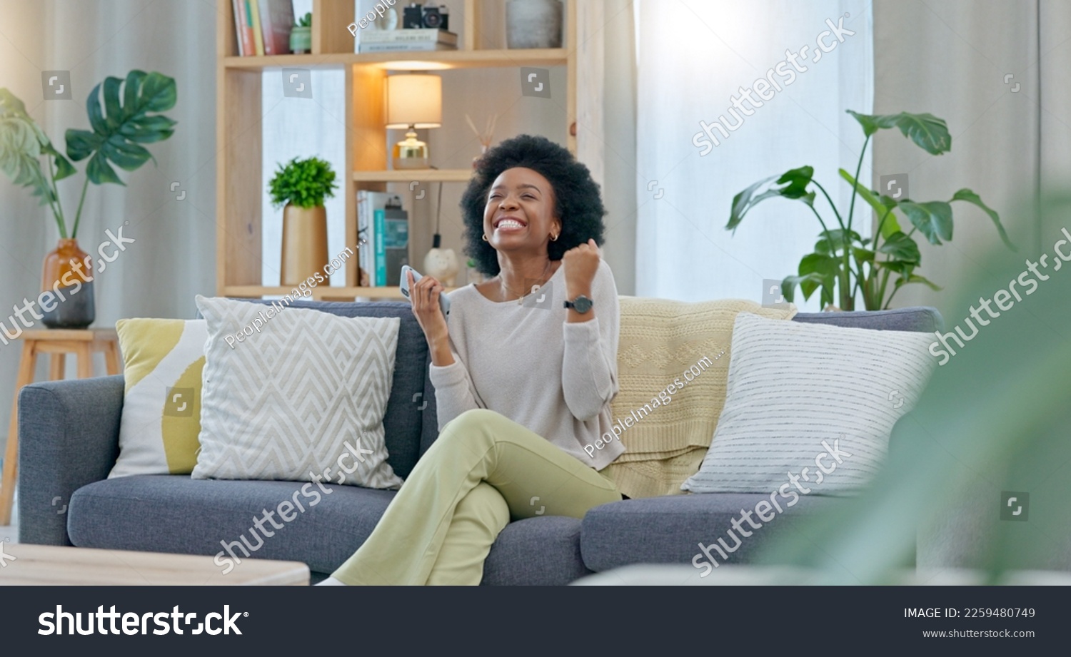 African woman celebrating a new job while sitting at home on a couch. A young females loan is approved via an email on her phone. A happy and excited lady cheering for a promotion on a sofa #2259480749