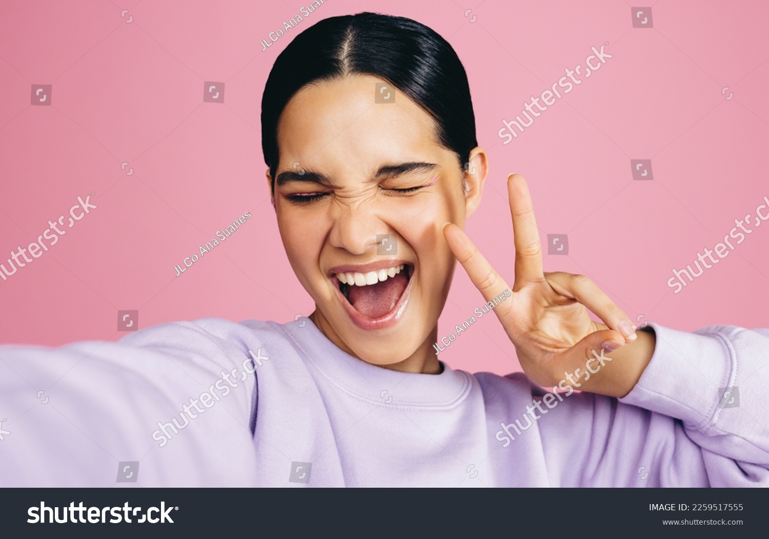 Excited young woman taking a selfie in a studio, she makes a peace sign with an expression of joy on her face. Woman having fun capturing her moments of self confidence and positivity. #2259517555