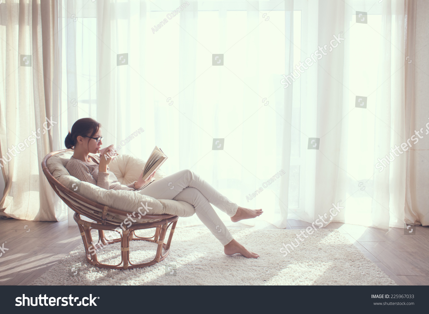 Young woman at home sitting on modern chair in front of window relaxing in her living room reading book and drinking coffee or tea #225967033