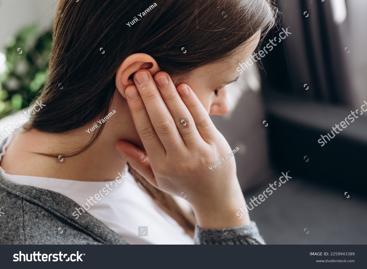 Close up of young brunette female holding painful ear, suddenly feeling strong ache. Unhealthy caucasian woman 20s suffering from painful otitis sitting on couch at home. Health problems concept #2259943389