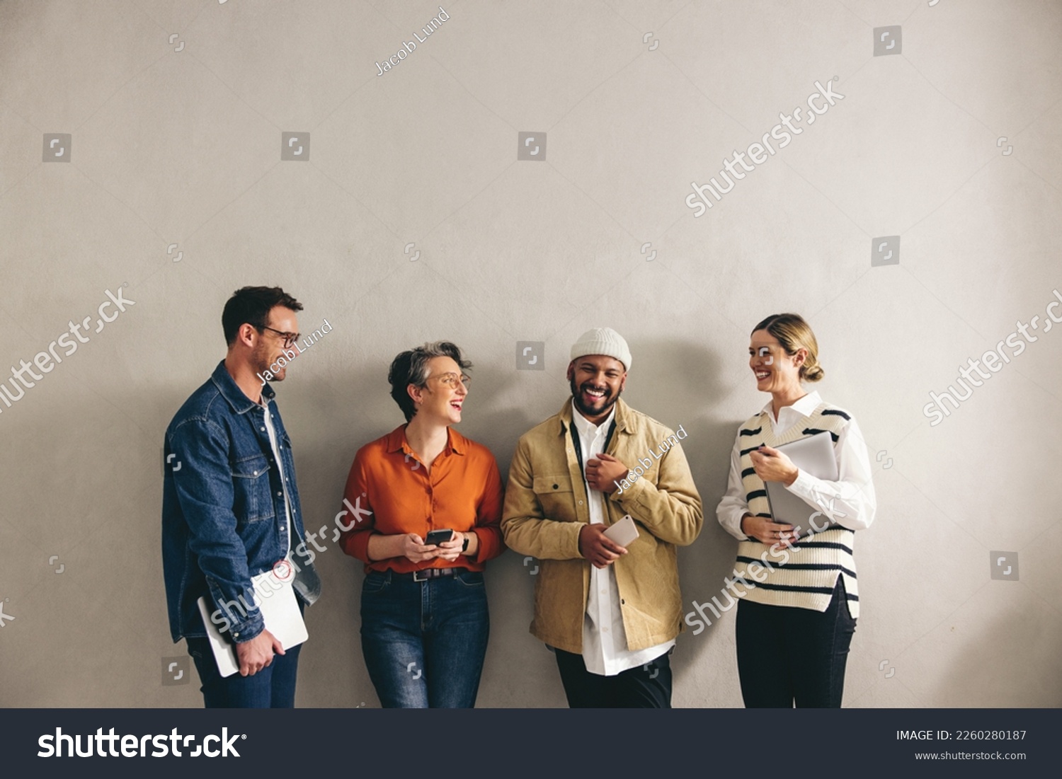 Happy businesspeople smiling cheerfully while waiting in line for an interview. Group of shortlisted job candidates holding different digital devices in a modern workplace. #2260280187