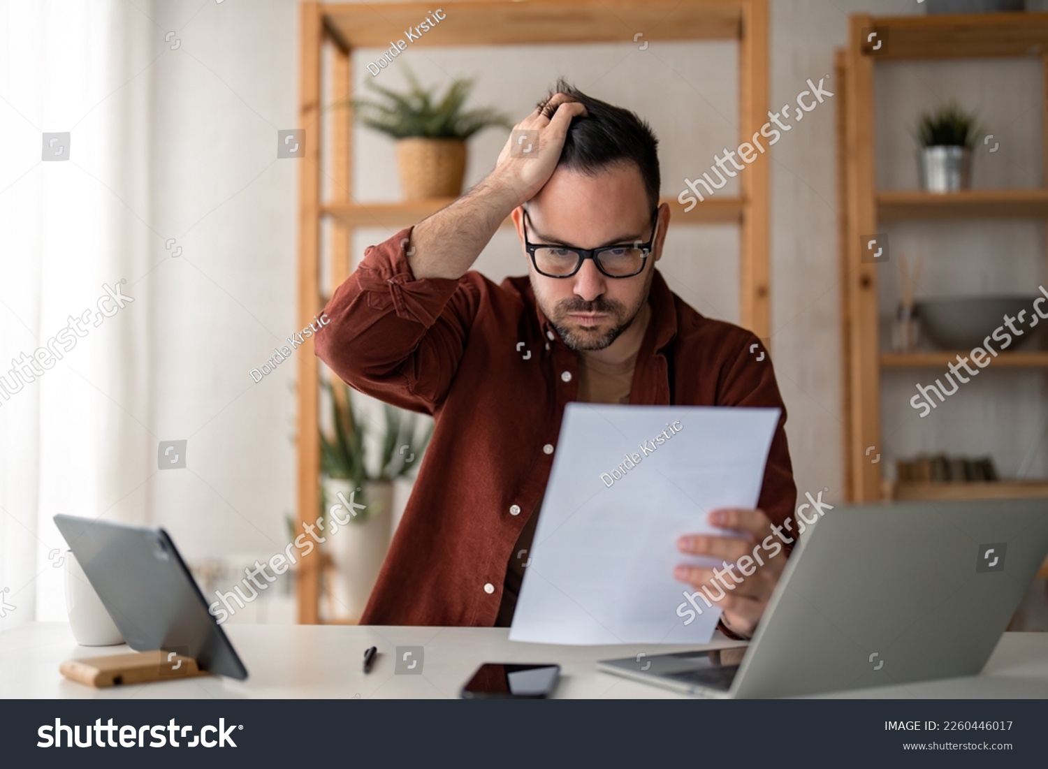 Overworked businessman at home office looking at paper document holding hand on his head feeling hopeless trying to find solution for given problem working too hard need break for better concentration #2260446017