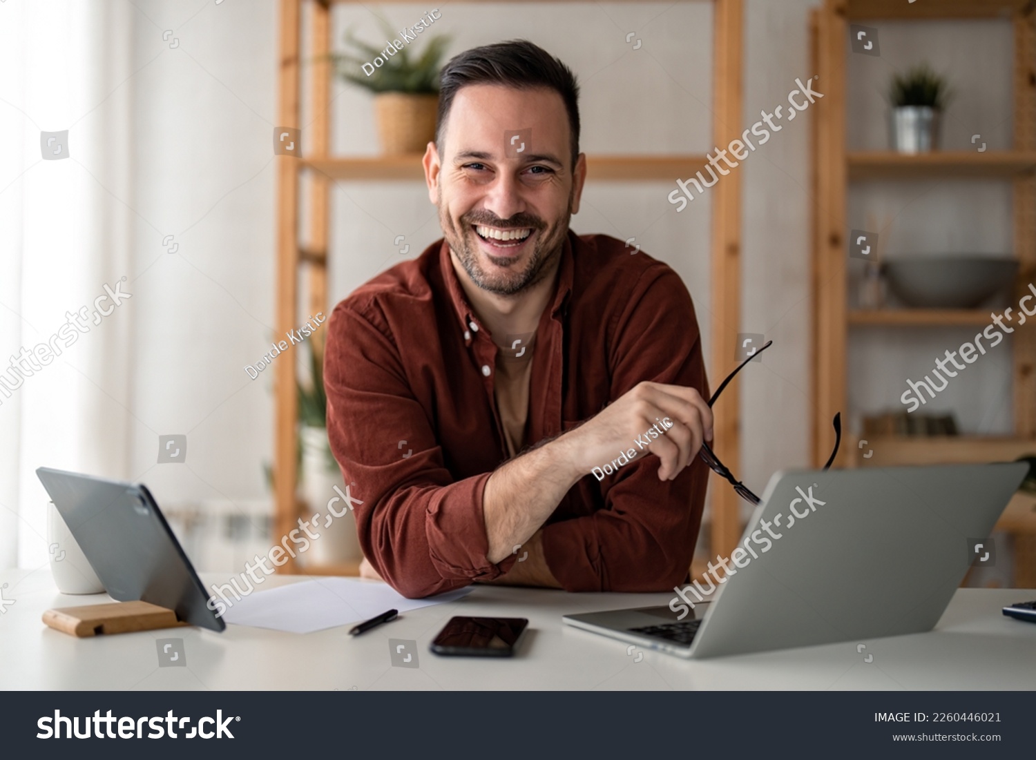 Smiling confident businessman looking at camera sitting at home office desk. Modern stylish corporate leader, successful manager or small business owner holding glasses posing for business portrait. #2260446021