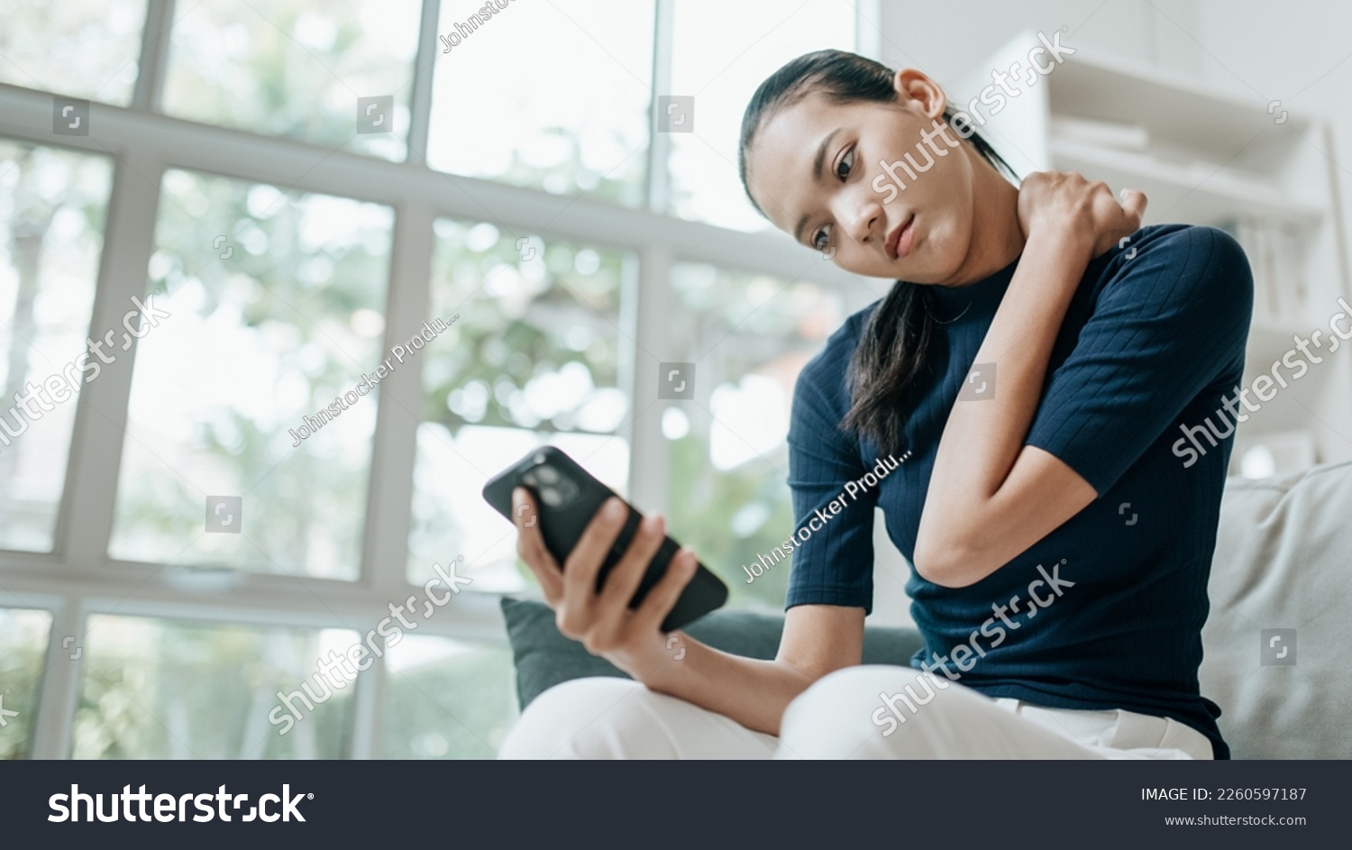 Portrait of Unhappy asian woman suffering from pain in the neck while sitting on the sofa and using mobile phone in the living room. Lifestyle, health care and medical concepts. #2260597187