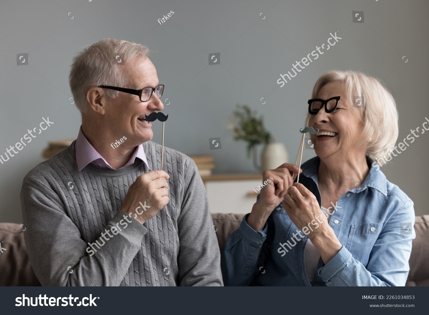 Happy joyful older couple of grandparents playing with photo shooting props, applying fake moustache and glasses on sticks to faces, having fun, smiling, laughing #2261034853