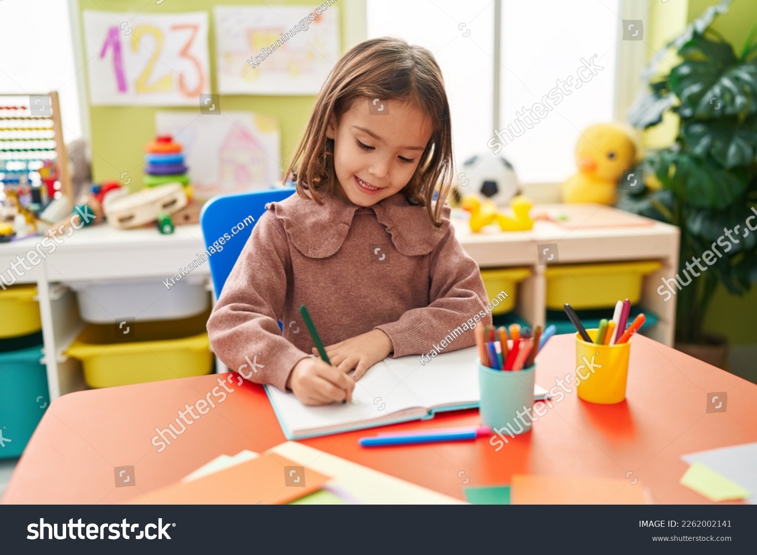 Adorable hispanic girl preschool student sitting on table writing on notebook at kindergarten #2262002141