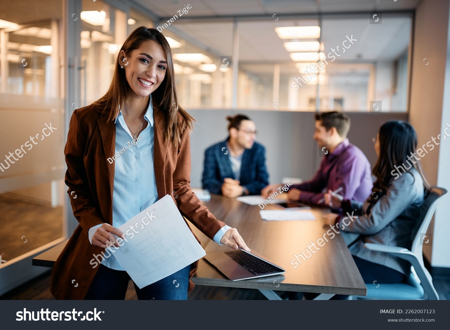 Portrait of young happy businesswoman during a meeting in the office looking at camera. Her colleagues are in the background.  #2262007123