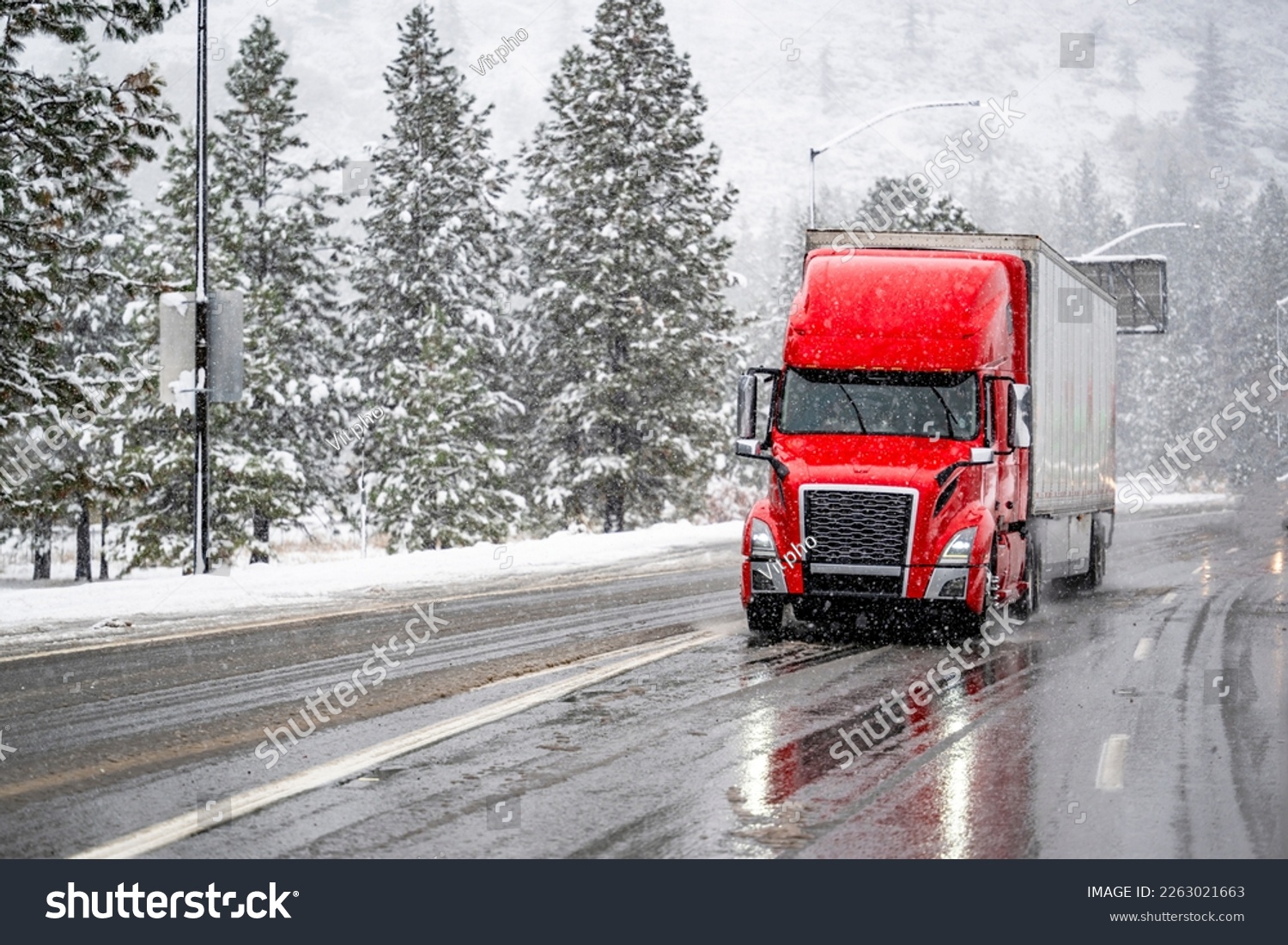 Red big rig commercial semi truck transporting cargo in dry van semi trailer running on the wet turning road with winter forest at snowing weather during a snow storm near Shasta Lake in California #2263021663