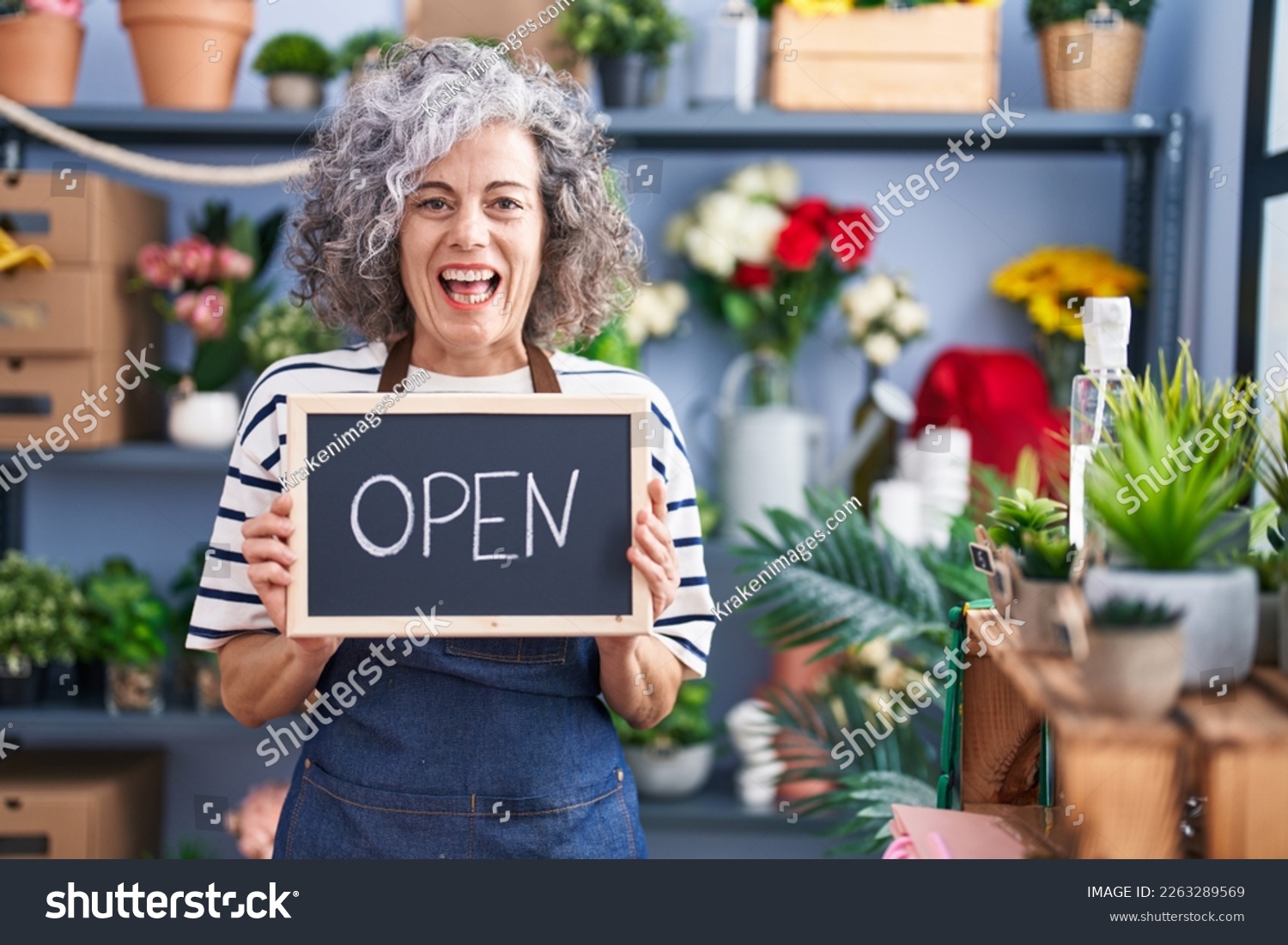 Middle age woman with grey hair working at florist with open sign smiling and laughing hard out loud because funny crazy joke.  #2263289569